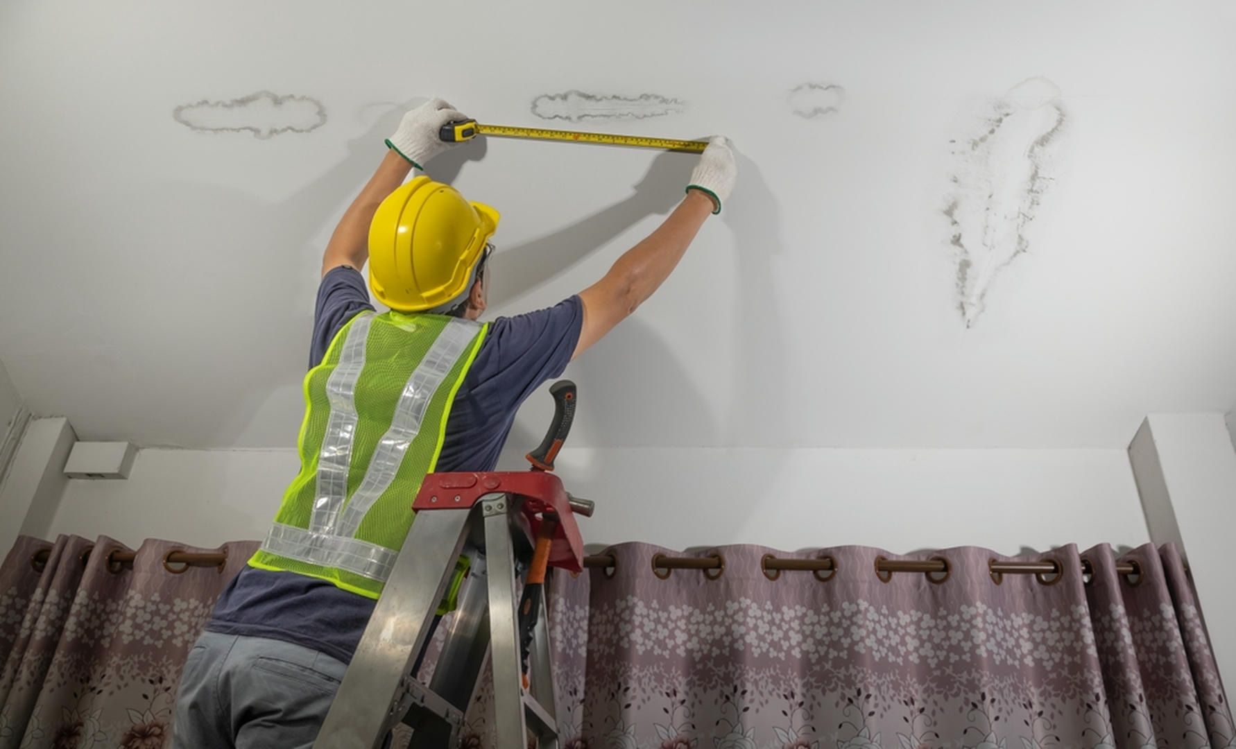 A man is measuring the ceiling with a tape measure while standing on a ladder.